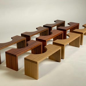 May include: A collection of wooden footstools in various shades of brown, from light oak to dark walnut. Each stool has a rectangular base and a curved top with a cutout, showcasing the natural wood grain.