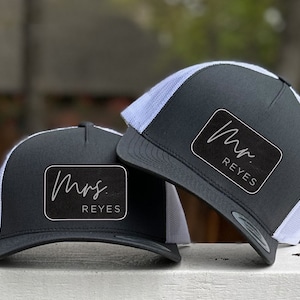 May include: Two black and white trucker hats with the names "Mr. Reyes" and "Mrs. Reyes" in a cursive font on a black patch. The hats have a mesh back and a curved brim.