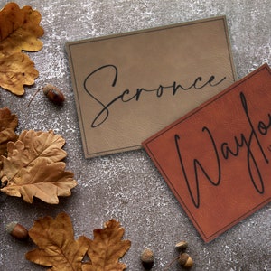 Pu&ograve; includere: Due etichette in pelle marrone con scrittura nera. L'etichetta superiore dice "Scronce" e l'etichetta inferiore dice "Waylon Wade". Le etichette sono su uno sfondo grigio con foglie di quercia marroni e ghiande.