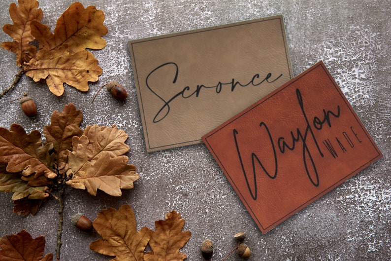 Pu&ograve; includere: Due etichette in pelle marrone con scrittura nera. L'etichetta superiore dice "Scronce" e l'etichetta inferiore dice "Waylon Wade". Le etichette sono su uno sfondo grigio con foglie di quercia marroni e ghiande.