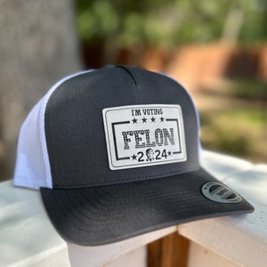 May include: A black and white trucker hat with a white mesh back. The hat has a white patch on the front that says "I'm Voting Felon *2024*" with a small image of Donald Trump.
