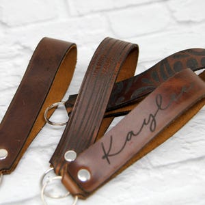 May include: Three brown leather keychains with silver key rings. One keychain has the name "Kayle" in a cursive font. The leather has a distressed look, and one keychain has a floral pattern.