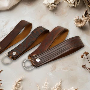 May include: Four brown leather keychains with silver key rings. The keychains have different designs, including a solid brown, a patterned design, and a striped design. Each keychain is secured with a silver rivet.