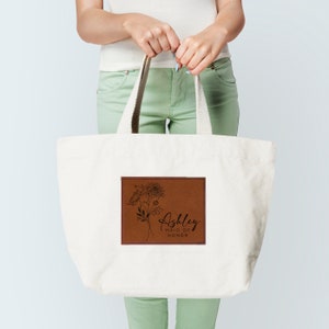 May include: A white canvas tote bag with brown leather patch featuring a floral design and the text "Ashley Maid of Honor".