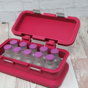 May include: An open, bright pink case with a hinged lid, containing several small, clear bottles with purple caps. The case is on a light wood-grain surface. The case has a rectangular shape with rounded corners.