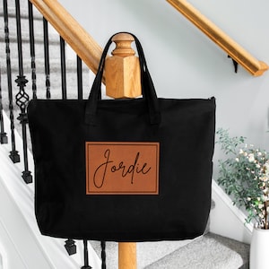 May include: A black tote bag with a brown leather patch featuring the name "Jordie" in a cursive font. The bag has two handles and a zipper closure. The bag is displayed on a wooden post near a staircase.