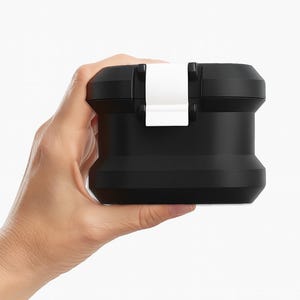 May include: A black, rectangular container with a white clasp is held in a hand. The container has a rounded base and a flat top. The clasp is centered on the top and appears to be made of plastic.
