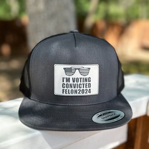 May include: A black trucker hat with a white patch that says "I'M VOTING CONVICTED FELON2024" and features a pair of sunglasses with an American flag design.