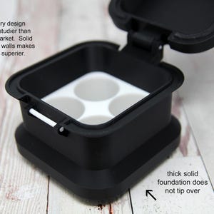 May include: Black square container with a hinged lid, featuring a white interior with four circular compartments. The text on the image says "Our proprietary design is significantly studier than others on the market. Solid base and thicker walls makes our cases far superior."