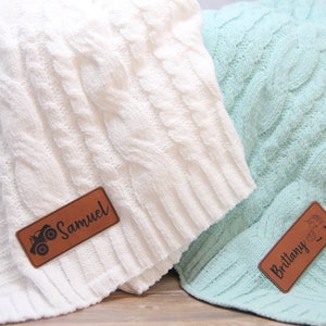 May include: Two white and light blue cable knit blankets with personalized leather labels. The white blanket has a brown label with a small truck graphic and the name "Samuel". The light blue blanket has a brown label with a small flower graphic and the name "Brittany".