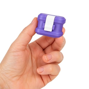 May include: A small, purple and white plastic object with a rectangular shape is held in a hand. The object has a white clasp and a ridged design. The background is plain white.