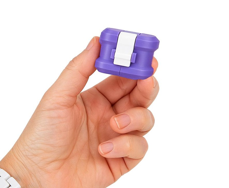 May include: A small, purple and white plastic object with a rectangular shape is held in a hand. The object has a white clasp and a ridged design. The background is plain white.