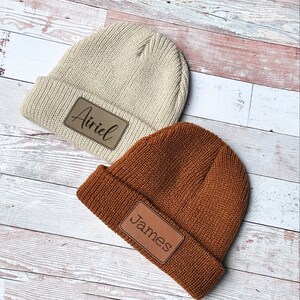 May include: Two knitted beanies with leather patches. The beanie on the left is beige with the name "Airiel" on the patch. The beanie on the right is brown with the name "James" on the patch.