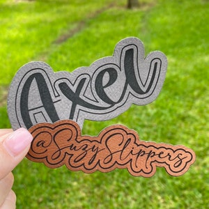 May include: Two custom-made name tags, one with the name "Axel" in black lettering on a gray background and the other with the text "Suzy Slippers" in black lettering on a copper background.