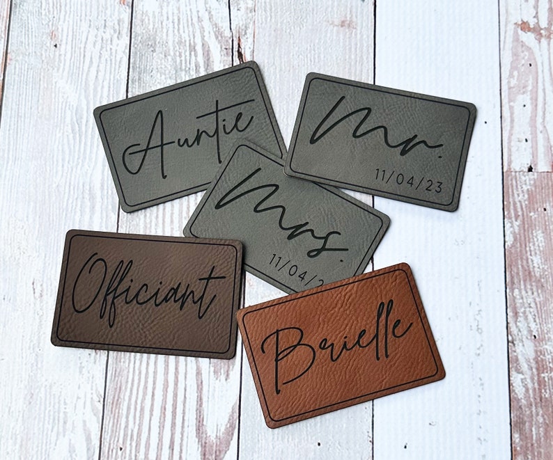 May include: Five brown and gray leather place cards with black script lettering. The cards read "Officiant", "Auntie", "Mr. 11/04/23", "Mrs. 11/04/23", and "Brielle".