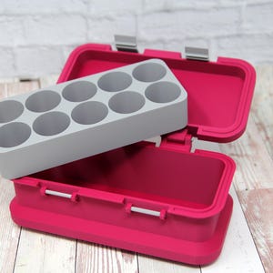 May include: A bright pink container with a gray insert featuring circular compartments. The container is open, revealing the interior and the insert. The container has silver hinges and latches. The background is a white brick wall and a wooden surface.