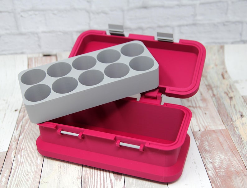 May include: A bright pink container with a gray insert featuring circular compartments. The container is open, revealing the interior and the insert. The container has silver hinges and latches. The background is a white brick wall and a wooden surface.