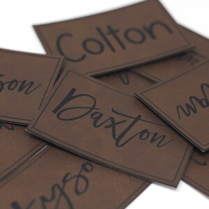 May include: A pile of brown leather name tags with black lettering. The names on the tags are: Dawson, Landon, Colton, Daxton, and  "Mr. Joel".