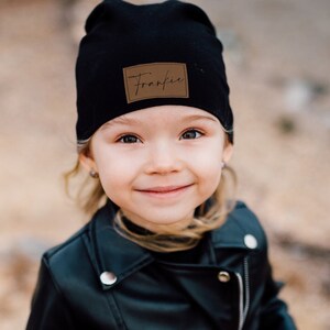 May include: A young girl wearing a black beanie with a brown leather patch that says "Frankie" on it. She is wearing a black leather jacket and has a smile on her face.
