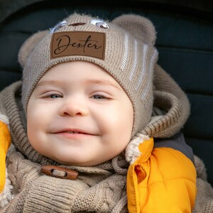 May include: A smiling baby wearing a beige knit hat with bear ears and a brown leather patch that reads "Dexter". The baby is also wearing a beige knit sweater and a yellow jacket.