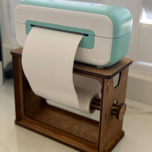 May include: A teal and white label printer on a wooden stand with a roll of white paper. The printer is on a white surface.