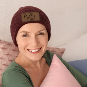 May include: A woman wearing a maroon beanie with a brown leather patch that says "faith over fear" is smiling and looking at the camera. She is sitting on a bed with pillows behind her.