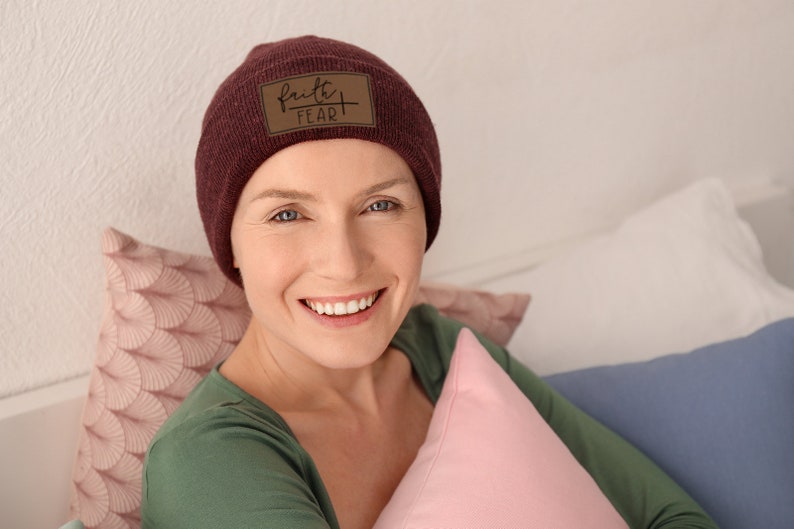 May include: A woman wearing a maroon beanie with a brown leather patch that says "faith over fear" is smiling and looking at the camera. She is sitting on a bed with pillows behind her.