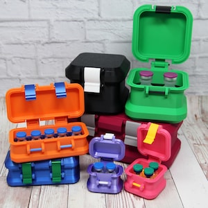 May include: A collection of colorful, small, hinged storage containers. The containers are in various colors, including blue, orange, green, pink, purple, and black. Some are open, revealing small vials inside. The containers have latches and are stacked.