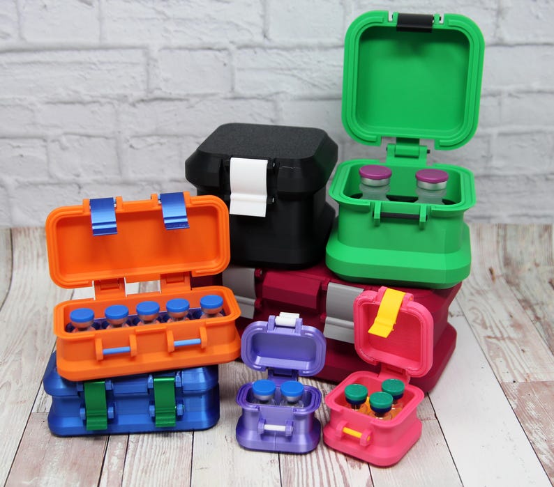 May include: A collection of colorful, small, hinged storage containers. The containers are in various colors, including blue, orange, green, pink, purple, and black. Some are open, revealing small vials inside. The containers have latches and are stacked.
