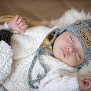 May include: A sleeping baby wearing a gray knitted hat with a brown leather patch that says "Jacob" on it. The baby is wearing a white knitted sweater and has a gray knitted tie.