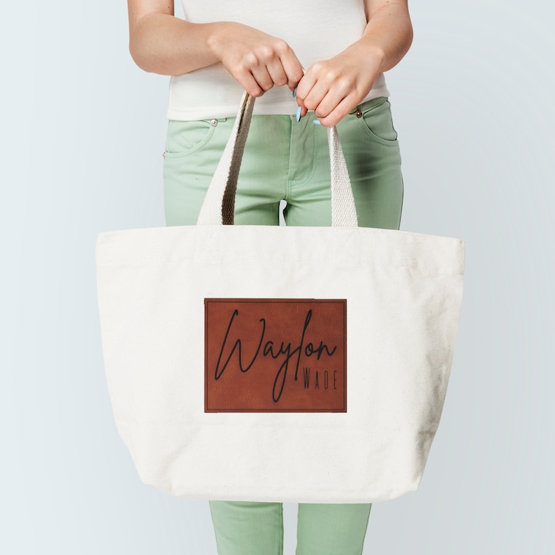 Pu&ograve; includere: Una borsa tote in tela bianca con una toppa in pelle marrone che dice "Waylon Wade" in scrittura nera.