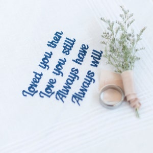 May include: White handkerchief with blue embroidered text that reads "Loved you then, Love you still, Always have, Always will". A small bouquet of white flowers and a silver ring are on top of the handkerchief.