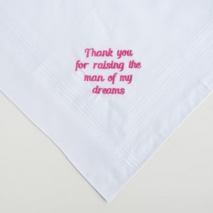Puede incluir: Pa&ntilde;uelo blanco con un mensaje bordado en rosa que dice: "Gracias por criar al hombre de mis sue&ntilde;os". El pa&ntilde;uelo tiene un borde texturizado y est&aacute; doblado en forma de tri&aacute;ngulo.