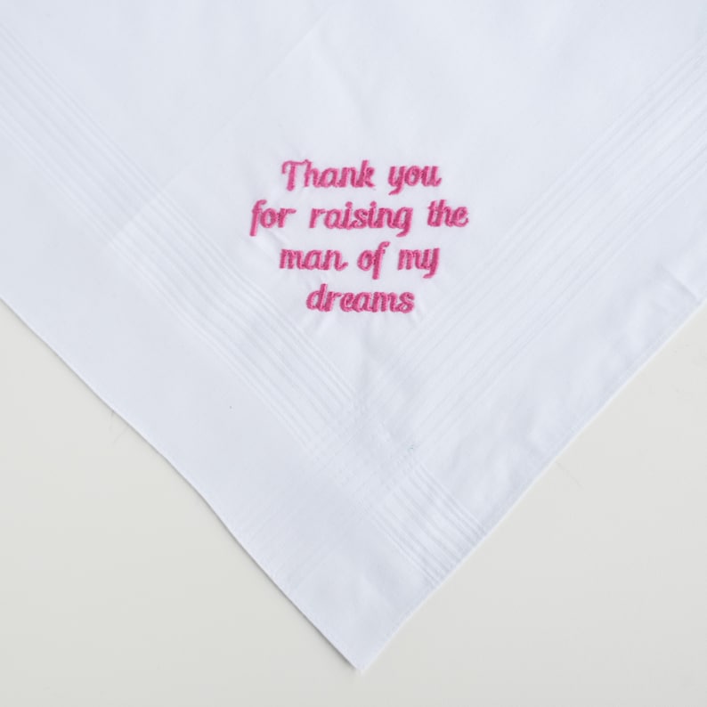 Puede incluir: Pa&ntilde;uelo blanco con un mensaje bordado en rosa que dice: "Gracias por criar al hombre de mis sue&ntilde;os". El pa&ntilde;uelo tiene un borde texturizado y est&aacute; doblado en forma de tri&aacute;ngulo.