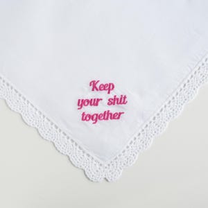 May include: White handkerchief with a delicate lace trim. The phrase "Keep your shit together" is embroidered in pink thread. The handkerchief is folded, showing a corner of the square fabric.