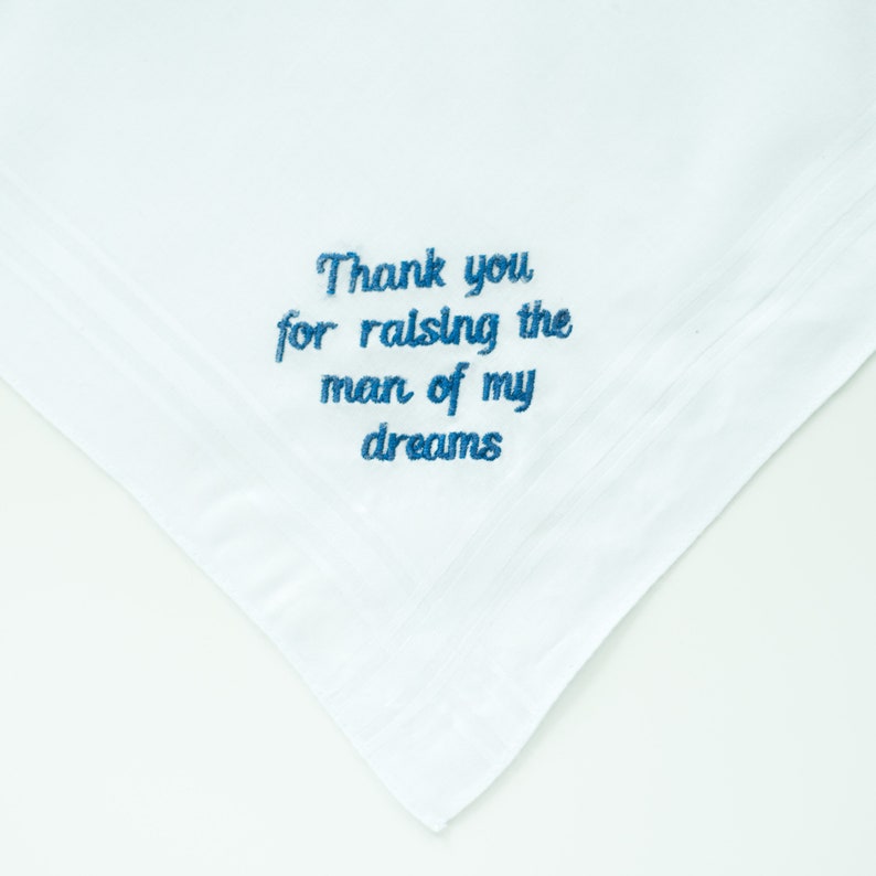 Puede incluir: Pa&ntilde;uelo blanco con texto bordado en azul que dice: "Thank you for raising the man of my dreams". El pa&ntilde;uelo est&aacute; doblado en una esquina y tiene un dise&ntilde;o sencillo y elegante.