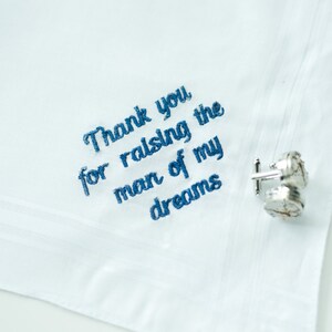 Puede incluir: Pa&ntilde;uelo blanco con texto azul bordado: "Thank you for raising the man of my dreams." Tambi&eacute;n se ven unos gemelos plateados con un dise&ntilde;o detallado.