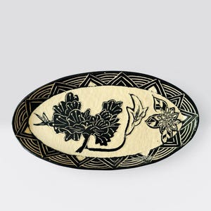 Pode incluir: Uma travessa oval de cerâmica com um centro creme e uma borda geométrica preta. O centro apresenta desenhos florais e de folhas pretas. A borda tem um padrão repetitivo de triângulos e linhas curvas.