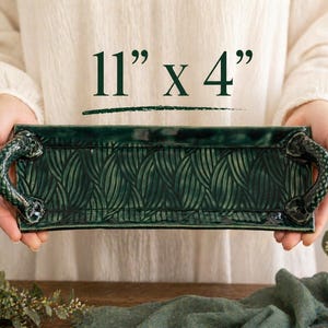 May include: A rectangular, dark green ceramic tray with leaf pattern and decorative handles. The tray measures 11 inches long by 4 inches wide. The handles are a darker shade of green. The tray is held by a person.