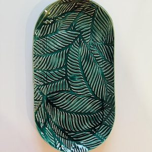 May include: An oval-shaped, teal ceramic serving platter with a raised leaf pattern. The platter has a glossy finish and a slightly raised edge. The leaf design is detailed and textured, creating a visually appealing surface.
