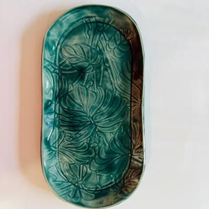 Puede incluir: Una bandeja ovalada de cerámica con esmalte turquesa. Presenta un diseño en relieve de hojas de monstera tropicales. El borde es marrón, agregando contraste. Esta bandeja decorativa es adecuada para servir alimentos o como pieza decorativa, midiendo unos 30 cm de largo.