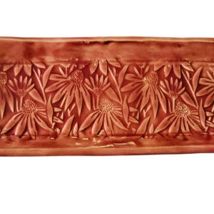 Puede incluir: Una bandeja rectangular de cerámica con un diseño floral. La bandeja es de color marrón rojizo con un patrón floral en relieve de flores y hojas estilizadas. Las asas son de color marrón oscuro y rojo rosado.