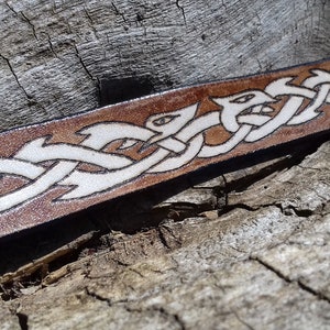 May include: A brown leather bracelet with a white Celtic knot design. The bracelet has a metal clasp.