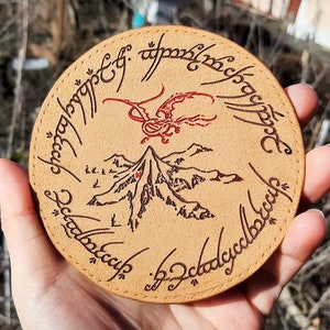 May include: A round, brown leather coaster with a red dragon and a mountain scene. The inscription around the edge reads "One Ring to rule them all, One Ring to find them, One Ring to bring them all and in the darkness bind them."