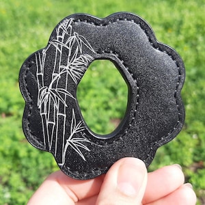 Puede incluir: Anillo de cuero negro con un diseño de bambú blanco. El anillo tiene un agujero en el centro.