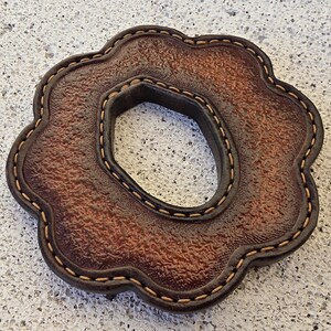 May include: A brown, flower-shaped leather coaster with a textured surface and a central hexagonal cutout. The edges are finished with a dark brown border and light tan stitching that follows the scalloped contour of the piece.