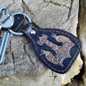 May include: A black leather keychain with a brown embossed design of a hammer and other symbols. The keychain is attached to a silver key ring with three keys.