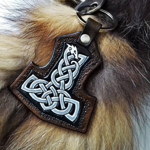 May include: A brown leather keychain with a silver Mjolnir hammer design. The hammer is surrounded by a Celtic knot design. The keychain is attached to a silver key ring.