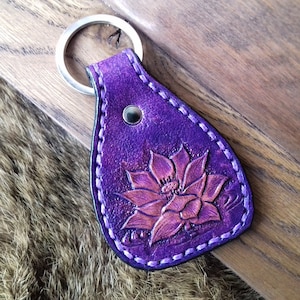May include: Purple leather keychain with a detailed embossed flower design. The keychain has a silver metal ring.