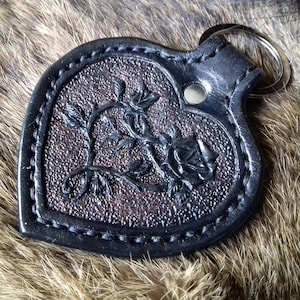 May include: A black leather heart-shaped keychain with a detailed rose design. The keychain has a silver metal ring.